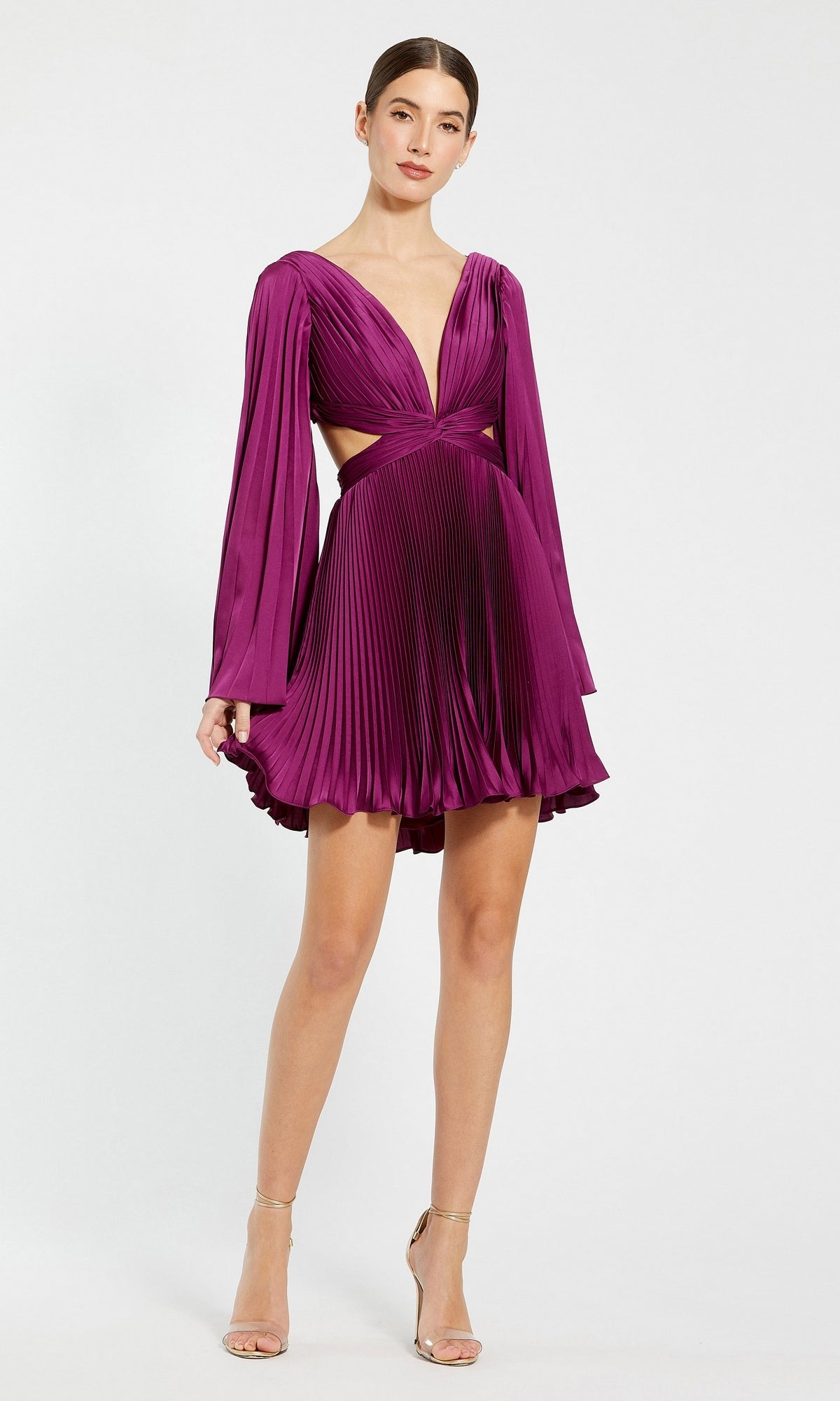 A woman models the Mac Duggal Long Sleeve Pleated Short Party Dress 27379 in deep purple, featuring long sleeves, a deep V-neckline, side cutouts, and pleats—styled with strappy heels for a chic semi-formal look against a white backdrop.
